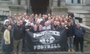 The friends of friar football take a group photo on the front steps of city hall in Providence with a Providence Football banner in front
