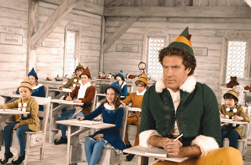 Buddy from the movie Elf sits at a tiny desk in a schoolroom with other elves