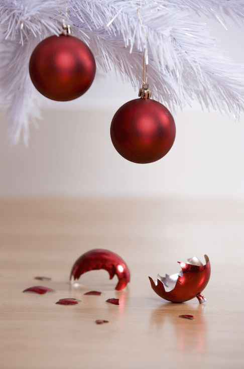 two red orb ornaments dangle precariously from a bottom branch of a christmas tree; on the ground lies a broken ornament