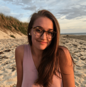 Michaela Campbell sits on the beach and smiles at the camera. She is wearing a pink tank top and glasses