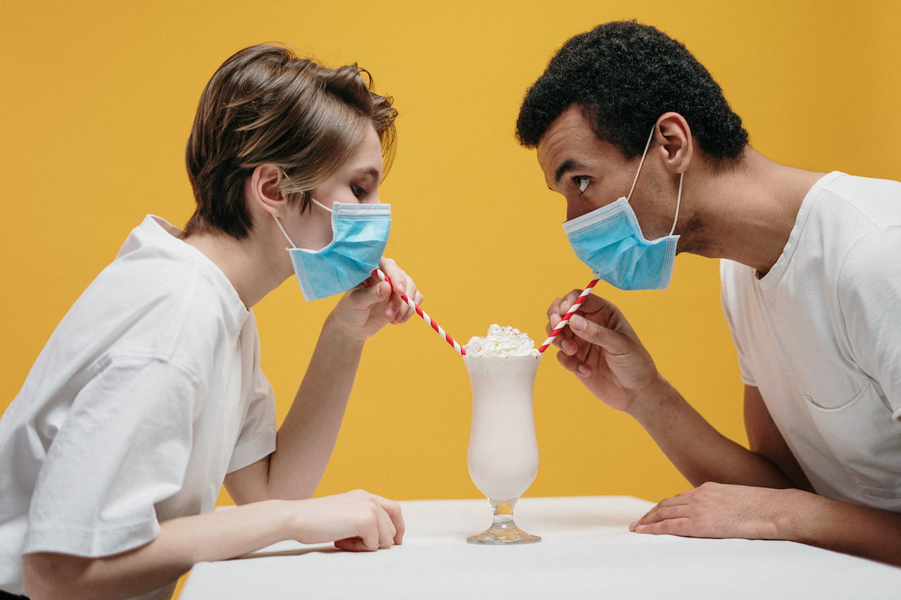 Two people attempting to share a milkshake while wearing surgical masks